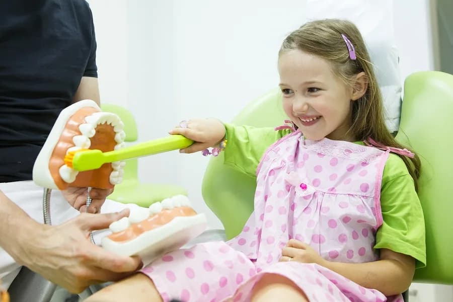 A smiling young girl in a dental chair practicing brushing on a large teeth model with an oversized toothbrush